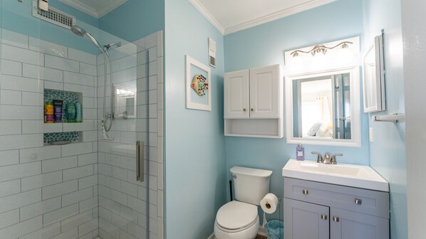 Ein Badezimmer mit einer Duschkabine aus Glas, weißen Fliesen, einer Toilette, einem Waschbecken mit Spiegel und Schränken.