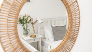 Un dormitorio con una cama blanca, una mesita de noche con lámpara, un florero con flores y un marco de espejo de mimbre.
