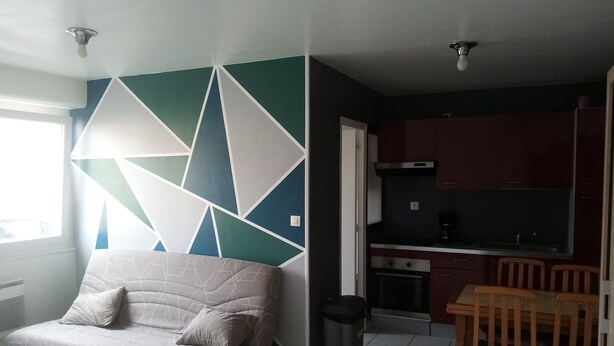 Un salon avec un canapé et un motif géométrique sur le mur, une cuisine avec des meubles sombres et un coin repas avec des chaises en bois.