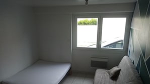 Une petite pièce, simplement aménagée, avec un lit simple, un canapé et une fenêtre donnant sur l’extérieur.