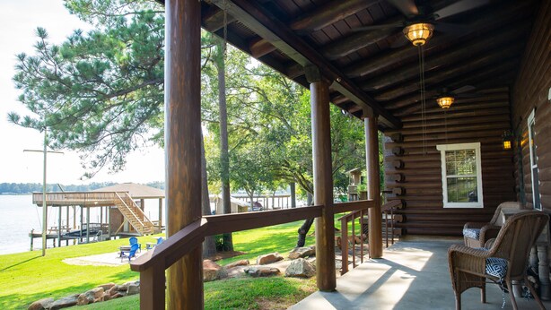 Una cabaña de madera con un porche que da a un lago, con terraza, asientos exteriores y una escalera que lleva a una zona cubierta.