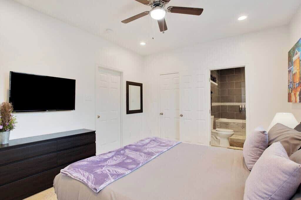 Une chambre à coucher avec un lit, un ventilateur de plafond, une télévision et une salle de bain visible à travers une porte ouverte.