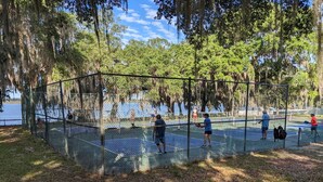 Una cancha de tenis con una cerca de malla metálica, rodeada de árboles y una masa de agua.