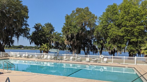 Piscina con sillas de descanso y sombrillas, rodeada de árboles y vista a un lago.