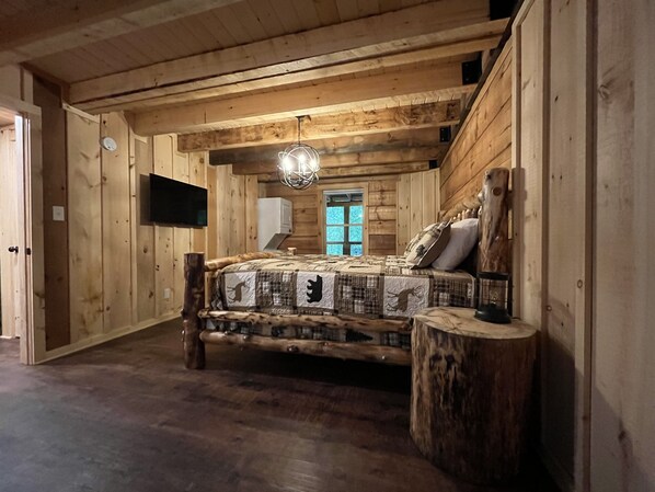 A log cabin bedroom with a bed, a mounted TV, a chandelier, and a window.