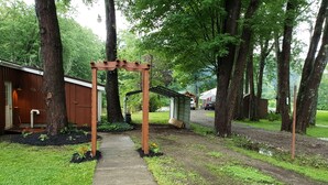 A wooden arbor leading to a covered porch, surrounded by trees and a gravel path. - Cozy Cottage (Ashville)