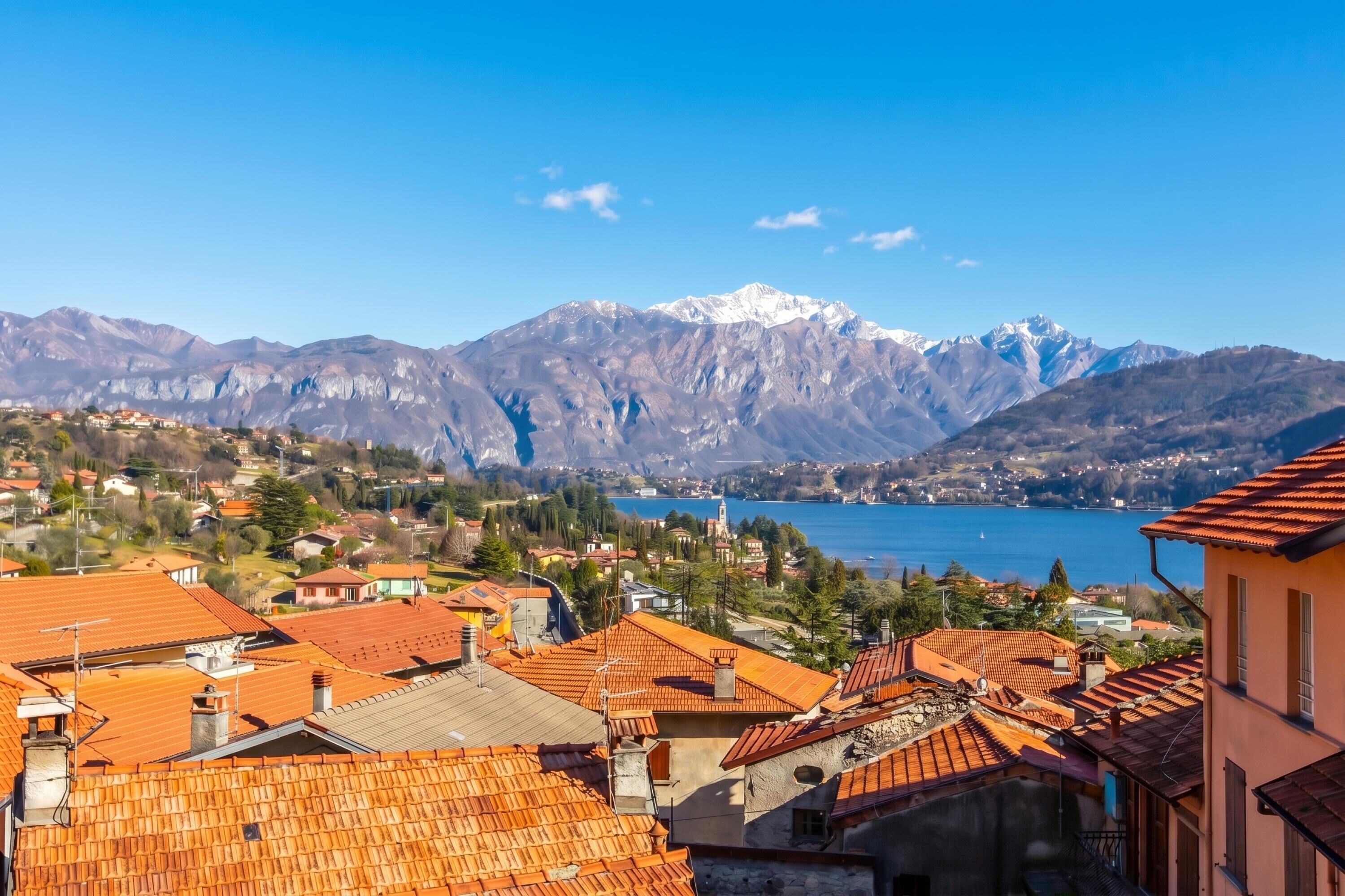 A town with terracotta-roofed buildings, a lake, and snow-capped mountains in the background.