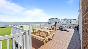 Een houten terras met picknicktafels en stoelen met uitzicht op een waterpartij, met een witte omheining en een gebouw op de achtergrond.