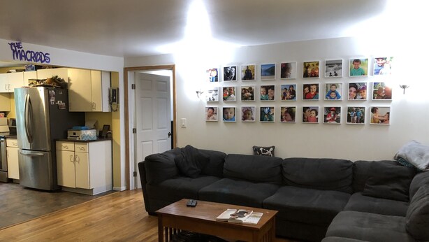 Un saló amb un sofà seccional gris fosc, una taula de centre de fusta, i una paret decorada amb fotografies emmarcades. Es veu, al fons, una cuina amb un frigorífic d'acer inoxidable i armaris.