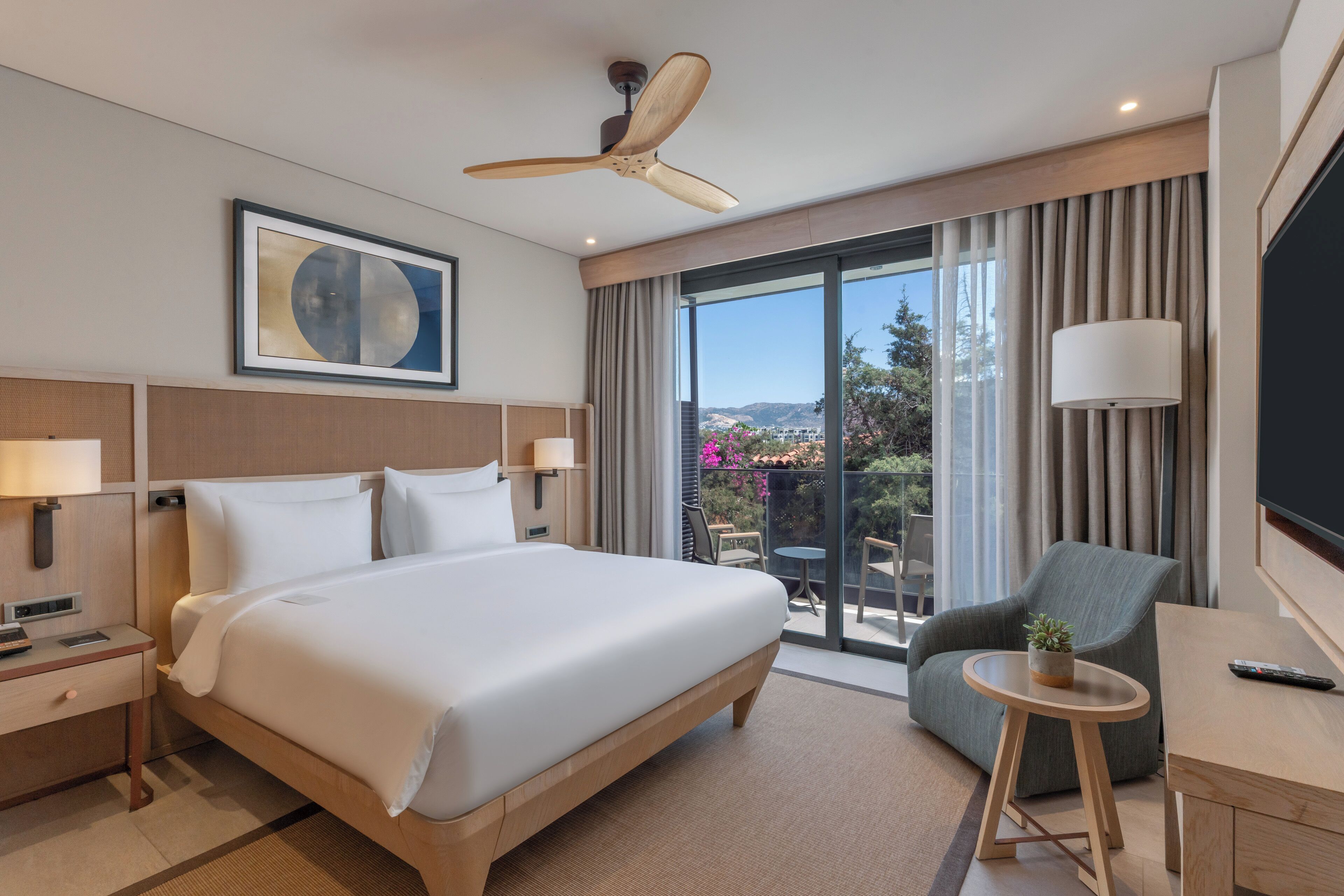 Habitación de hotel con una cama grande, un escritorio, una silla, un ventilador de techo, un televisor y un balcón con vistas.