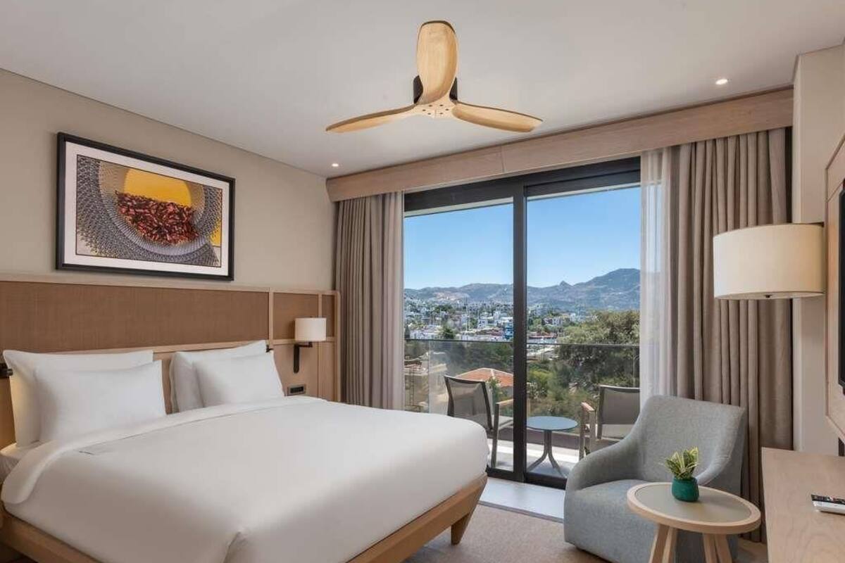 Une chambre d’hôtel avec un lit, un ventilateur de plafond, une vue sur la ville et un balcon équipé d’une table et de chaises.
