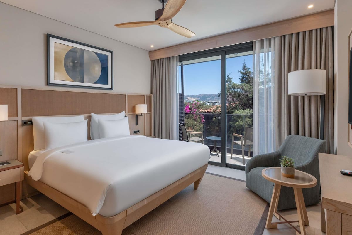 Habitación de hotel con una cama grande, un ventilador de techo, vistas al exterior, una zona de estar con mesa y sillas, y un televisor de pared.