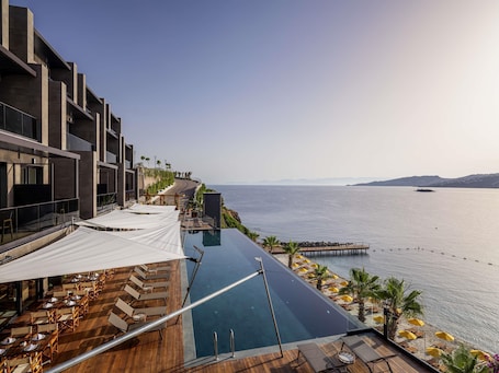 A modern hotel with a pool overlooking the sea, wooden deck seating, and a clear view of the coastline.
