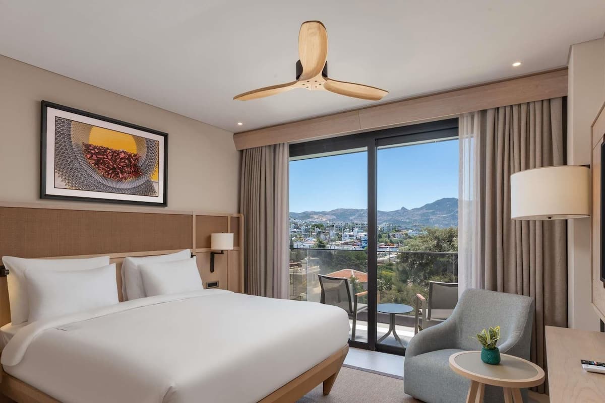 Habitación de hotel con una cama grande, un escritorio, una silla, una mesita con una planta, ventilador de techo y vista a la ciudad a través de amplias ventanas.