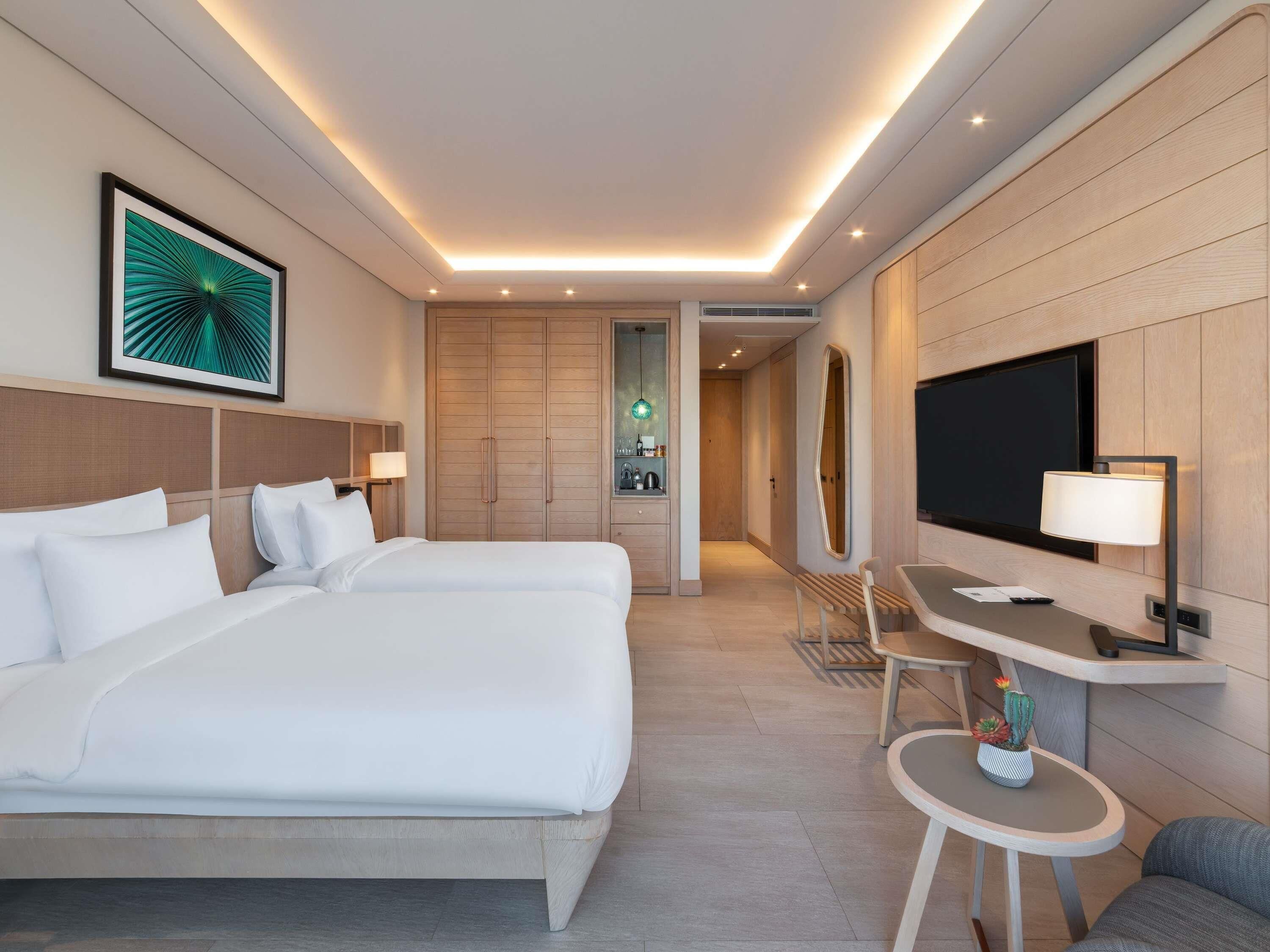 Habitación de hotel con una cama grande, un escritorio con televisor, un espejo y una mesita con una planta.