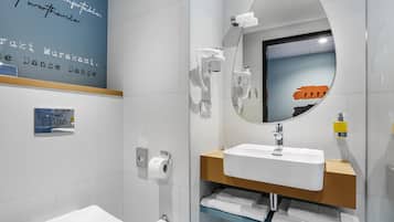 A modern bathroom with a white toilet, a sink with a mirror above it, and a towel rack with folded towels. There is a wall-mounted towel dispenser and a toilet paper holder.