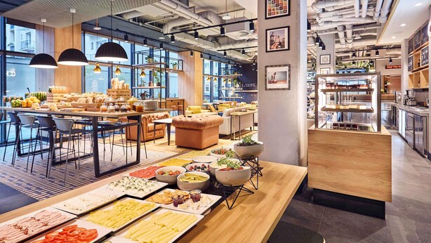 Un café moderno con una larga mesa compartida llena de diversos alimentos, disposición de asientos y un área de cocina visible.