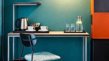 A room with teal walls, a desk with a kettle, cups, and glasses, a chair, a hanging lamp, and a black cabinet.