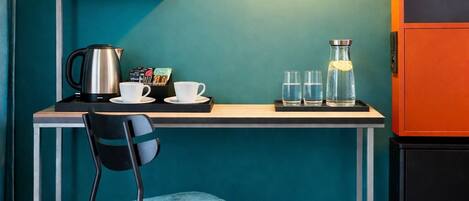 Een kamer met teal muren, een bureau met waterkoker, bekers en glazen, een stoel, een hanglamp en een zwart kastje.
