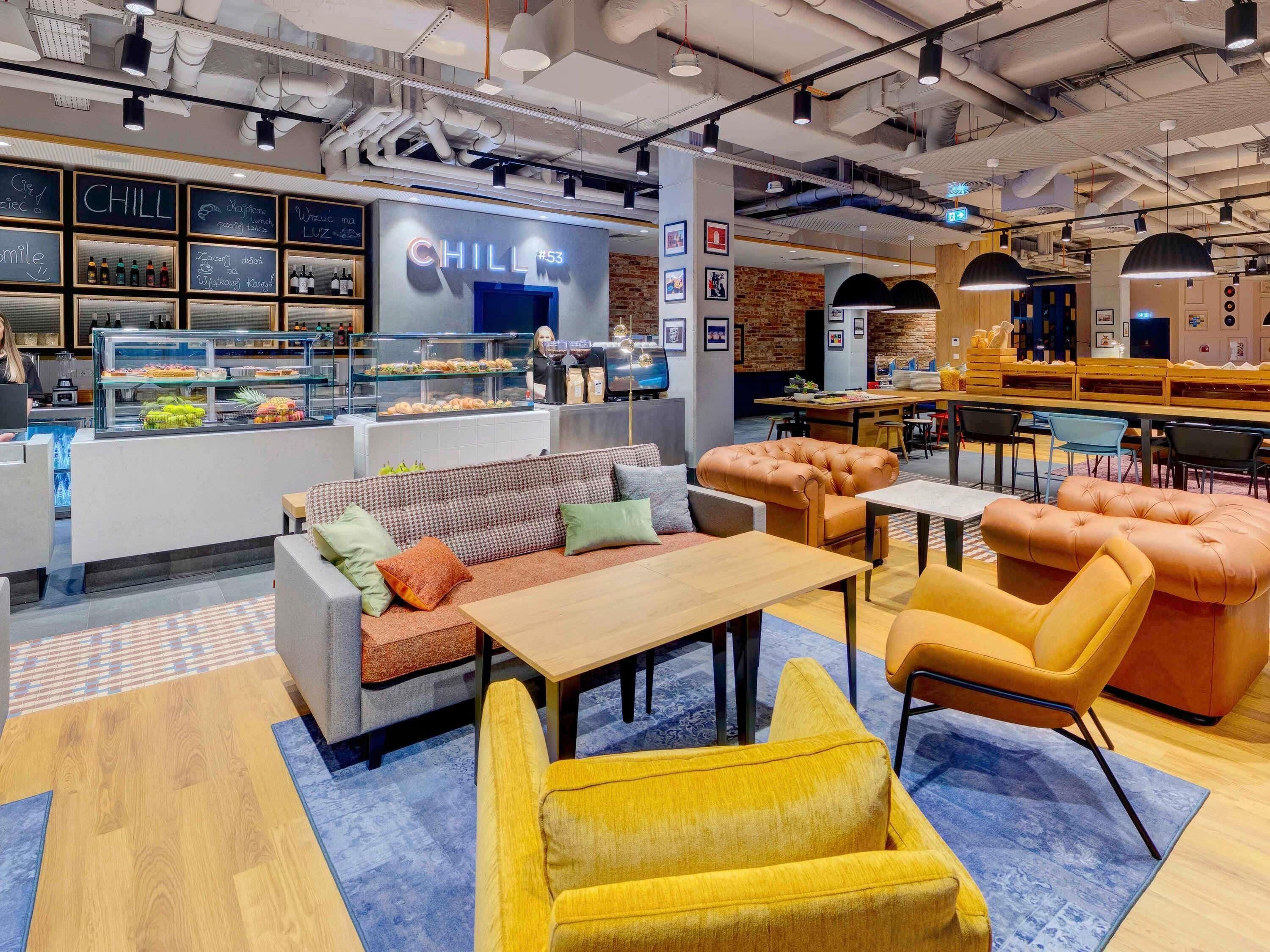 A modern cafe interior with a counter displaying beverages and pastries, seating arrangements including sofas and chairs, and a visible chalkboard menu.