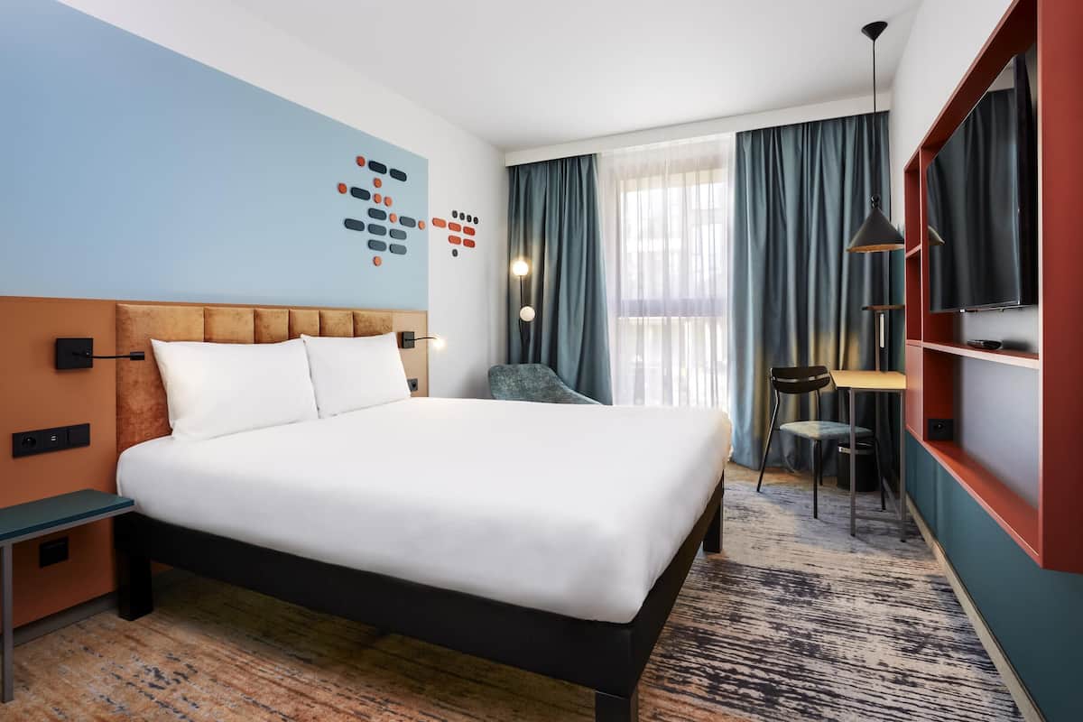 A hotel room with a bed, a desk, a chair, a TV, and a window with curtains.