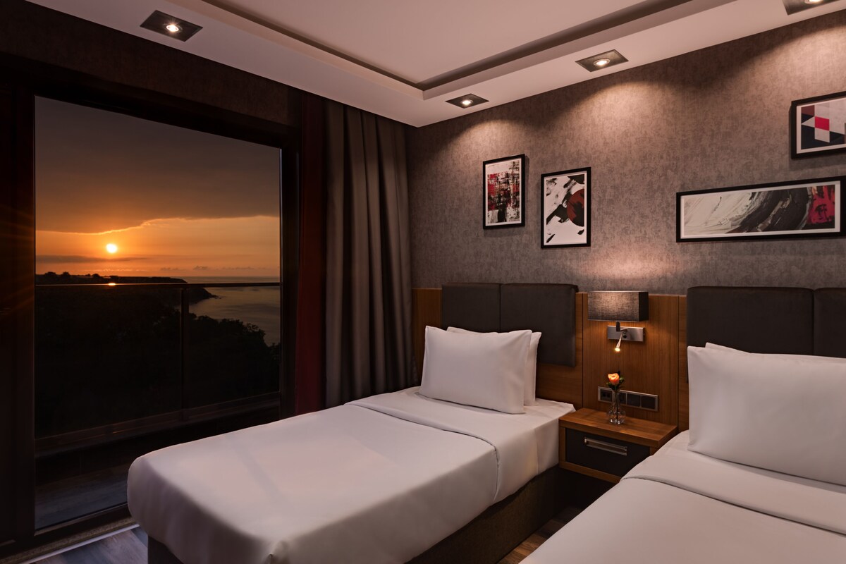 A hotel room with two beds, a large window with a sunset view, and artwork on the walls.