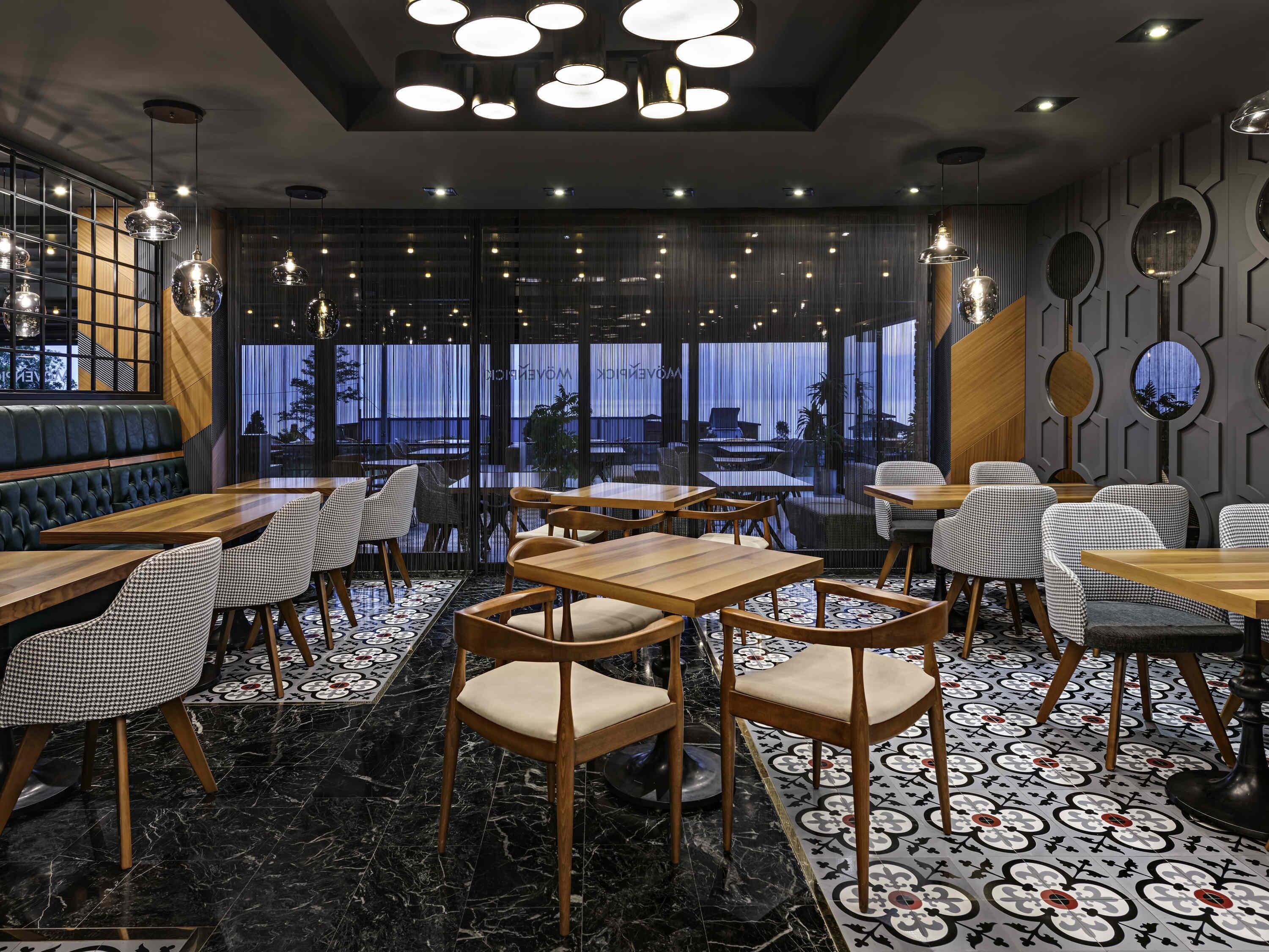 A modern restaurant interior with wooden tables and chairs, a patterned floor, and large windows.