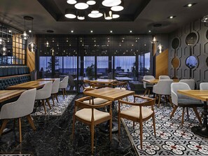 A modern restaurant interior with wooden tables and chairs, a patterned floor, and large windows. - Mövenpick Hotel Trabzon (Trabzon)