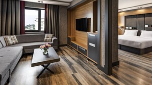 Una habitación de hotel moderna con sofá, una mesita con flores, televisor, minibar y un área de dormitorio separada con cama.