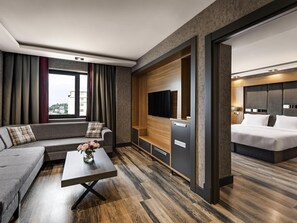 A modern hotel room with a sofa, a small table with flowers, a TV, a minibar, and a separate bedroom area with a bed. - Mövenpick Hotel Trabzon (Trabzon)