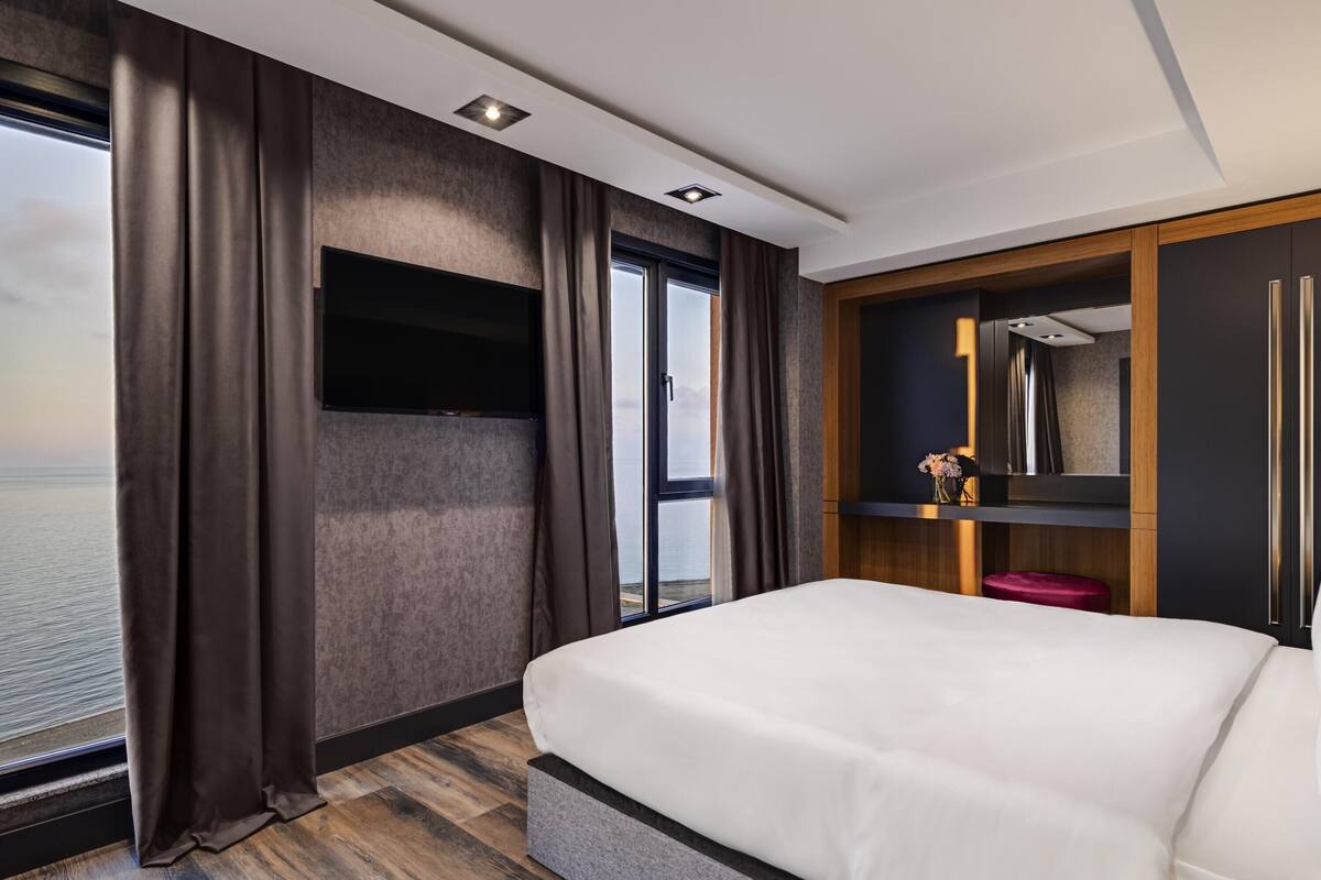 A hotel room with a large bed, a TV mounted on the wall, a wardrobe, and a view of the sea.
