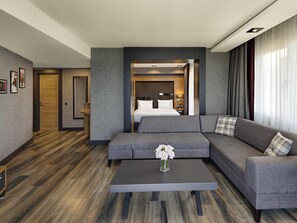 A modern hotel room with a grey sofa, a small table with a vase of flowers, a bed with white linens, and a wooden floor. - Mövenpick Hotel Trabzon (Trabzon)