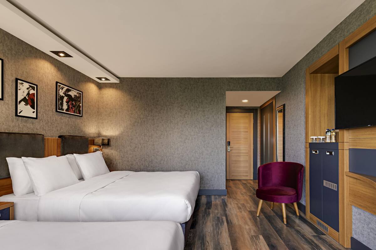 A hotel room with two beds, a wooden wardrobe, a television, and artwork on the wall.