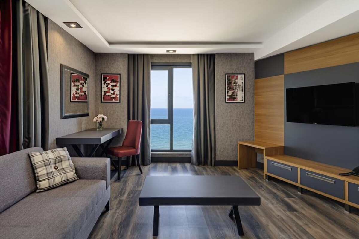A modern hotel room with a sofa, a small table, a chair, a TV, and a view of the sea.