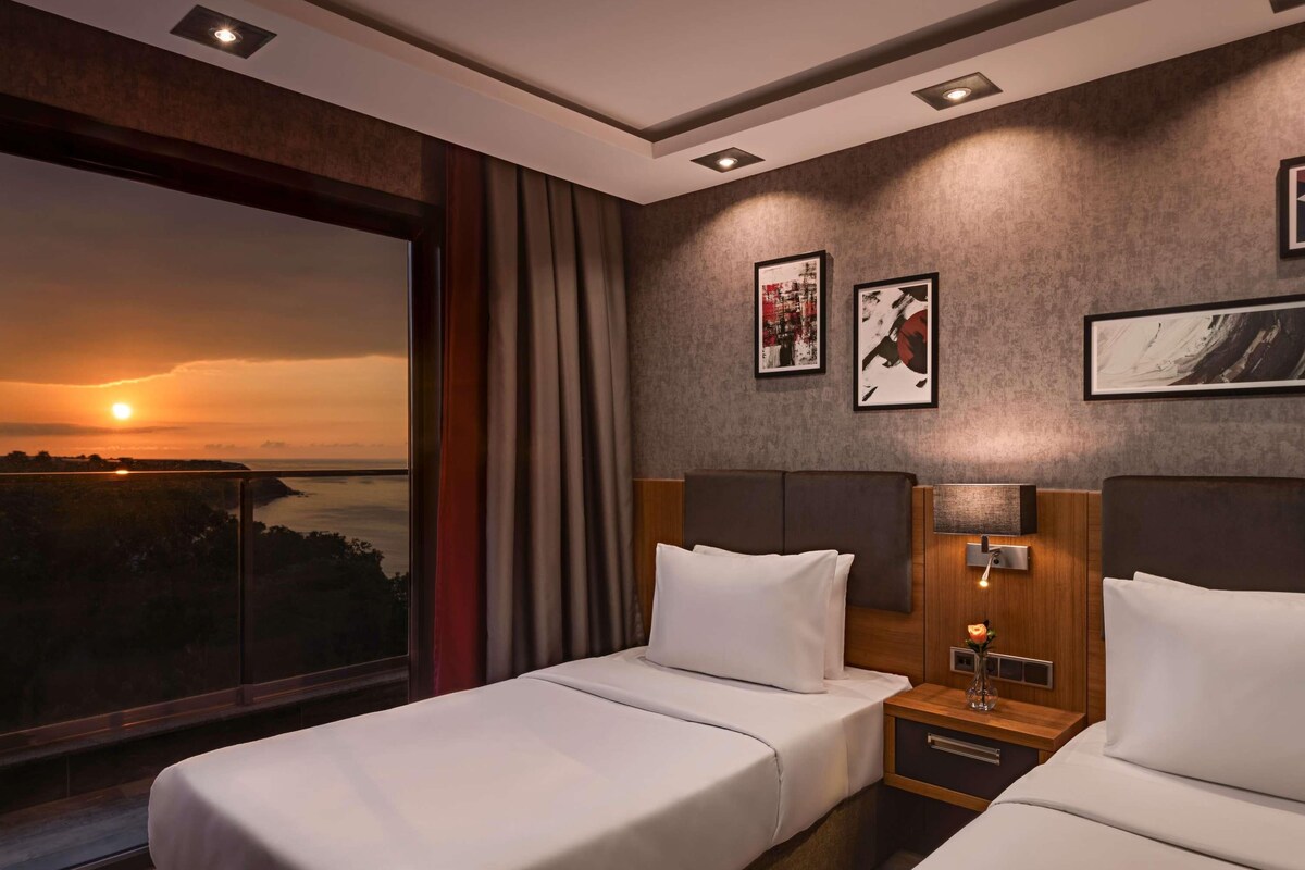 A hotel room with two beds, a large window with a sunset view, and artwork on the wall.