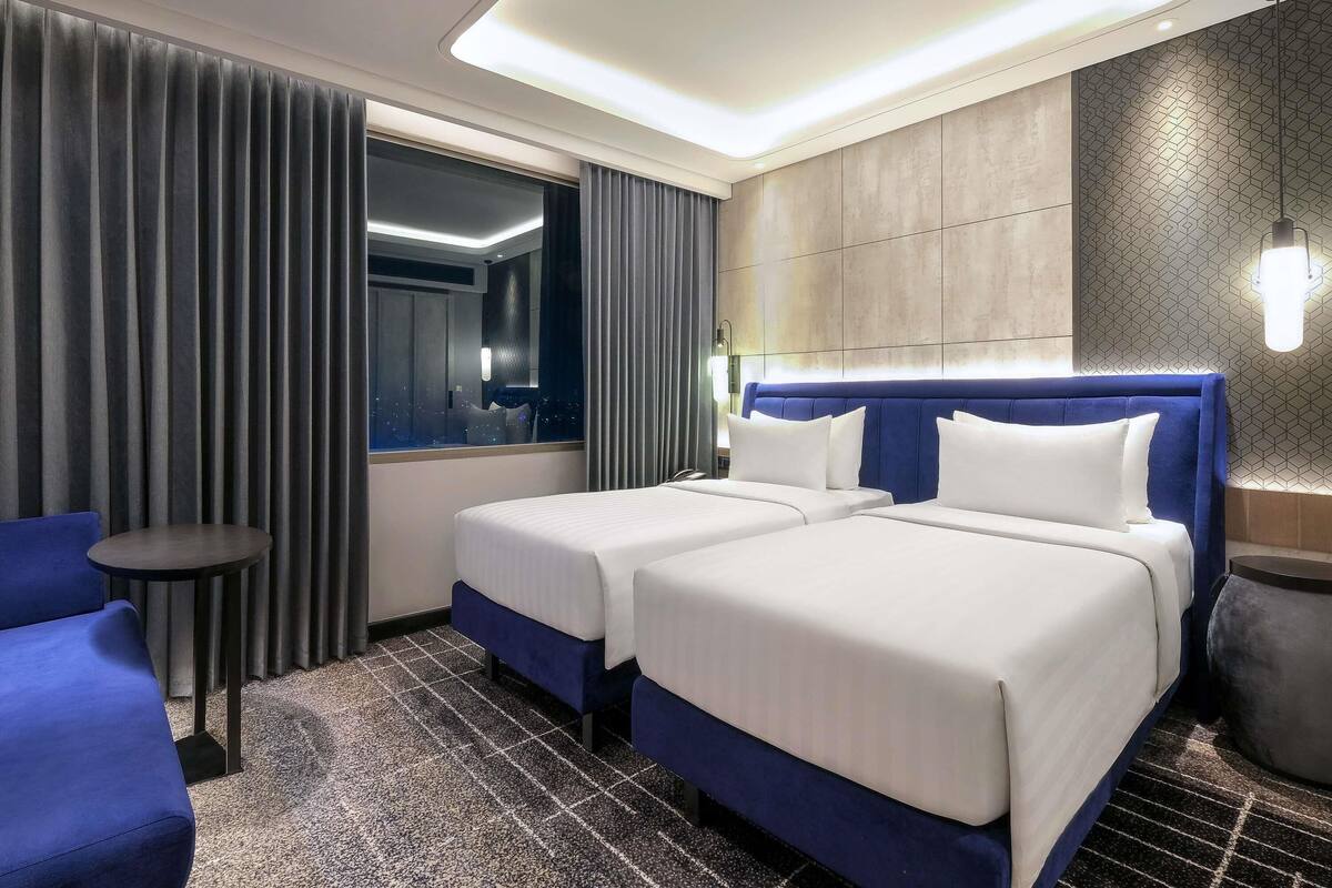 A hotel room with two beds, a blue chair, a small round table, and a large window with curtains.