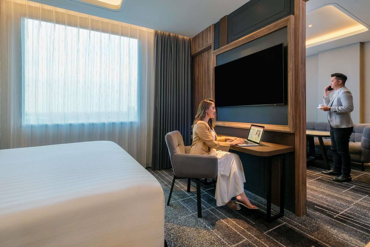 A hotel room with a bed, a desk with a laptop, a TV mounted on the wall, and a large window with curtains.