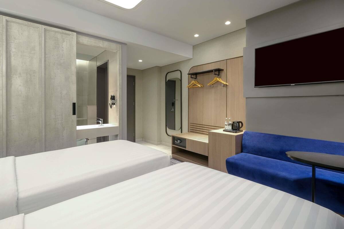 A modern hotel room with two beds, a large mirror, a wardrobe, a desk with a coffee maker, and a blue sofa.