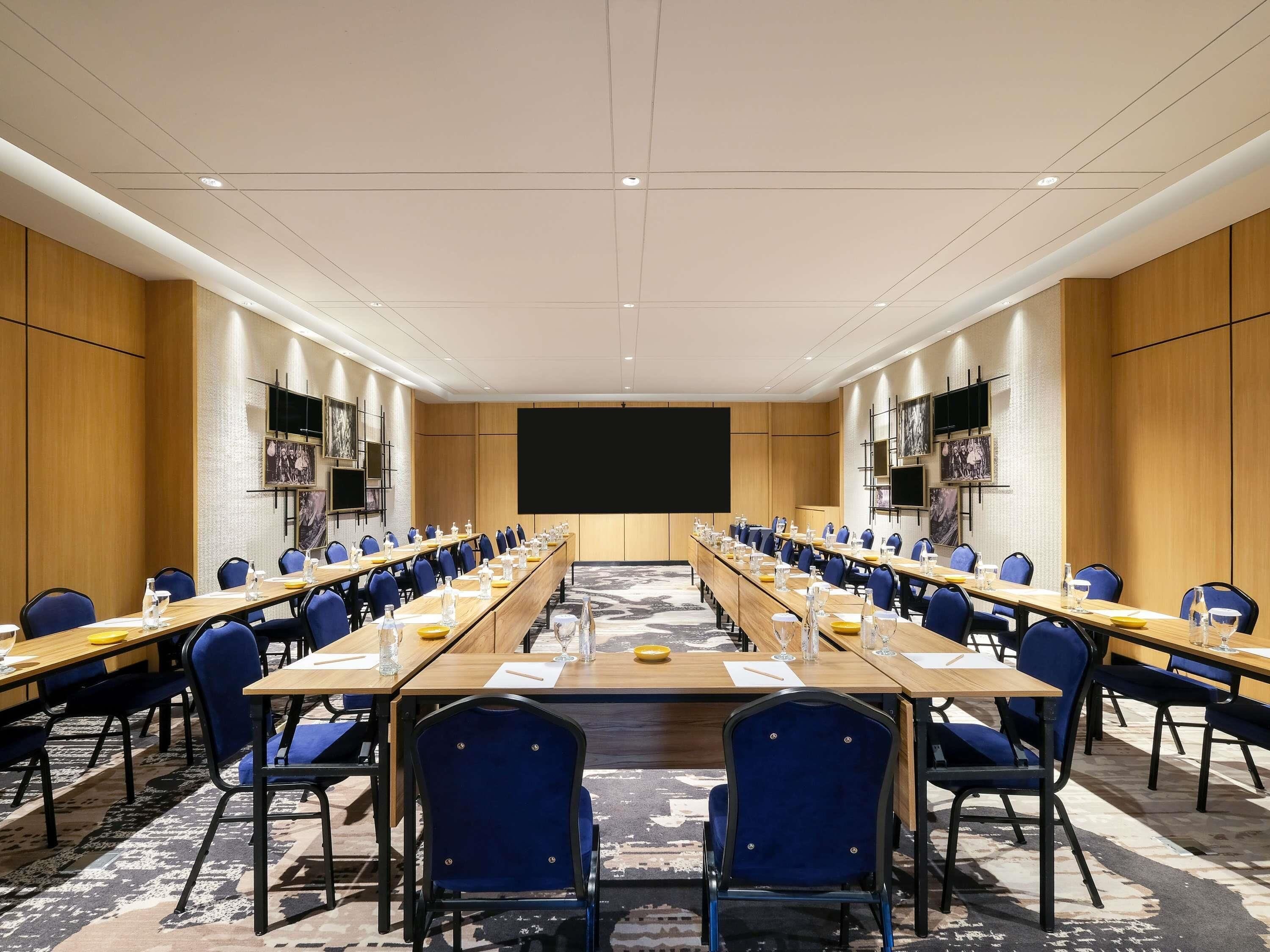 Un salón de conferencias con mesas largas, sillas azules y una pantalla grande.