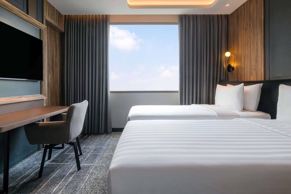 A hotel room with a desk, two beds, a TV, and a large window.