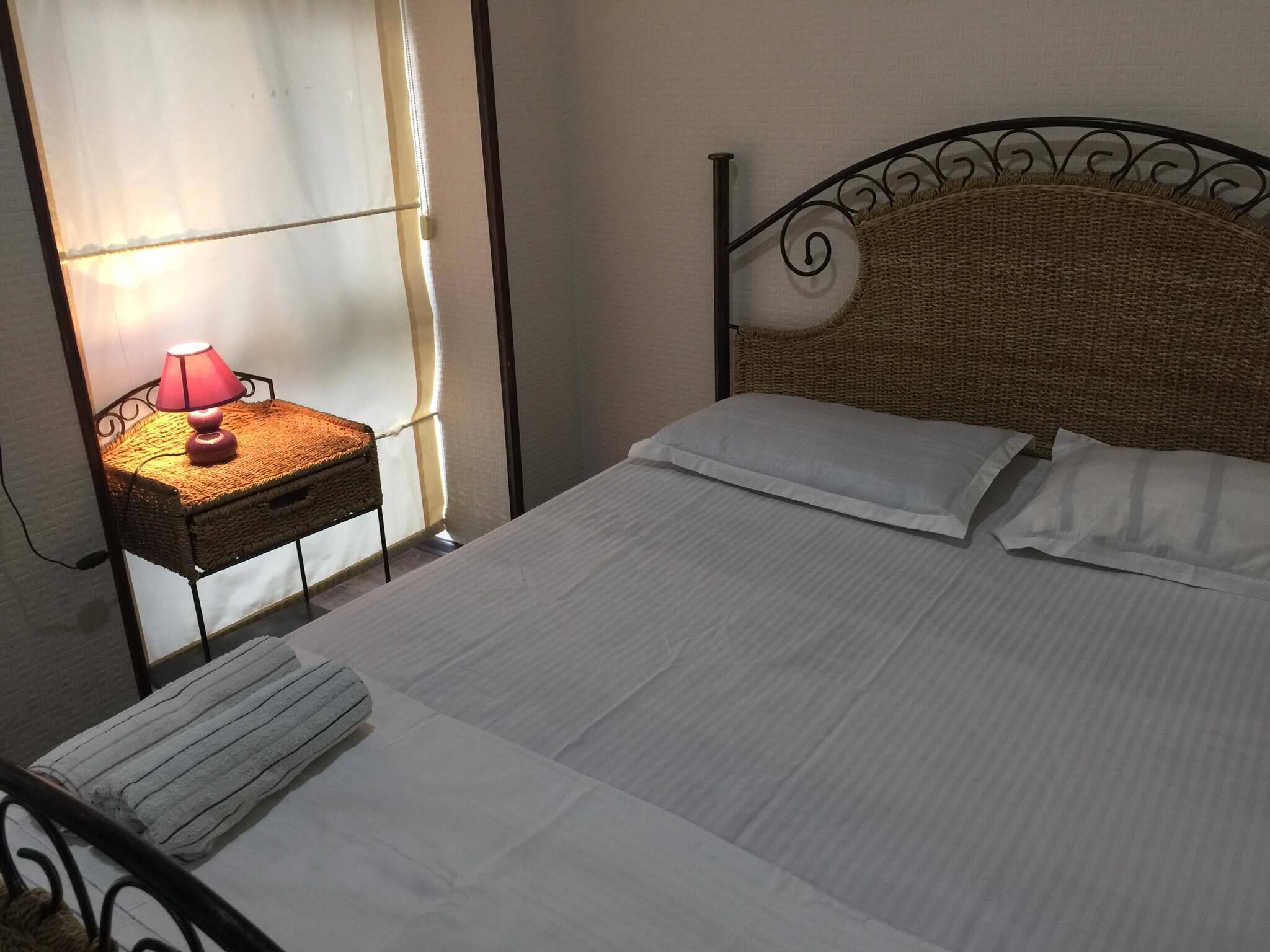 A neatly made bed with white linens, a bedside table with a lamp, and a window with sheer curtains.