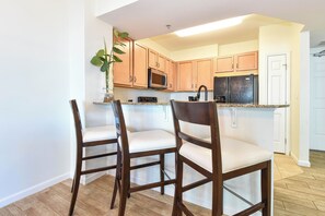Una cocina con armarios de madera, un microondas y un refrigerador. Hay dos sillas blancas con estructura de madera.
