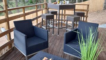 A covered outdoor seating area with wicker furniture and a table set for dining, surrounded by a wooden deck and trees.