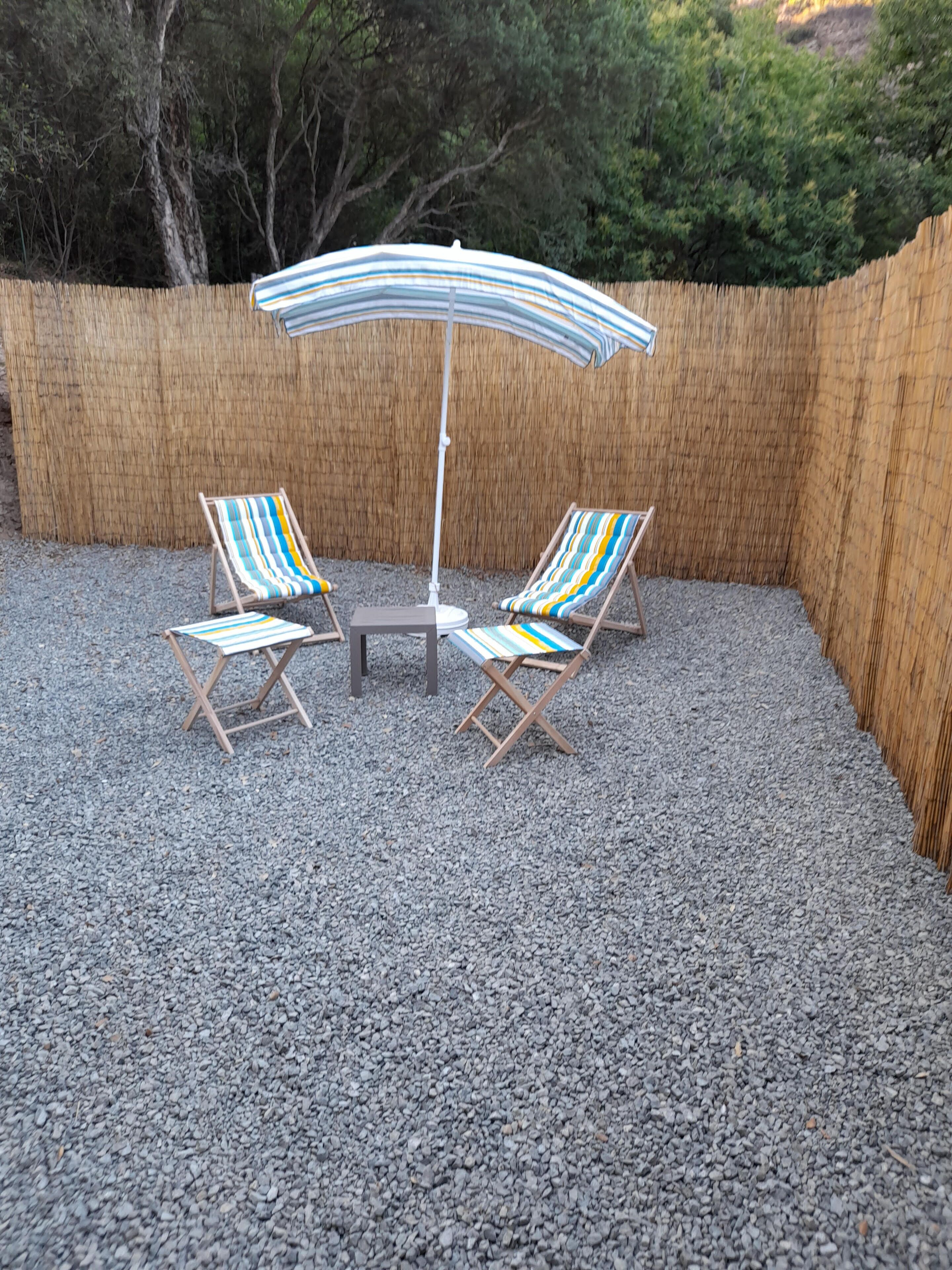Een terras met twee gestreepte lounge fauteuils en een tafel onder een gestreepte parasol, omringd door grind en een bamboe hek.