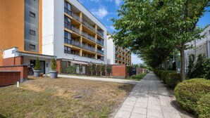 A multi-story residential building with balconies, a paved walkway, and a grassy area with potted plants.