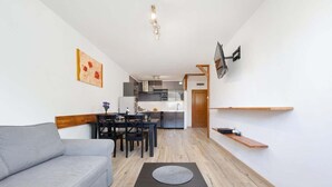A modern living room with a grey sofa, a dining table with chairs, a kitchen in the background, and a TV mounted on the wall.