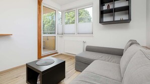 A modern living room with a grey sofa, a small black side table, a wooden-framed window, and a black shelving unit against the wall.