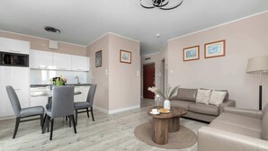 A modern living room with a sofa, a dining table with chairs, a kitchen area with cabinets, and a ceiling light fixture. - Apartment with Sea View | 200 m to the Beach, Balcony (Kolobrzeg)
