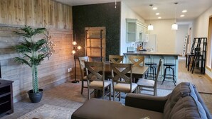 Ein modernes Wohnzimmer mit einer Wandpaneinung aus Holz, ein Essbereich mit Holztisch und -stühlen, eine Küche mit weißen Schränken und eine Bar mit Hockern.