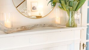 A fireplace with a white mantel, a round mirror, and a vase with white flowers. - Relaxing Coastal Inspired Cottage, short walk from the lake! (Ashtabula)